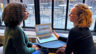 Young woman with short curly hair green sweater and glasses sits at small round desk with blond woman with dark shirt and glasses. On desk is a laptop with the word "Zotero" on the screen. Empty tables and chairs on patio are visible in background through glass wall. 