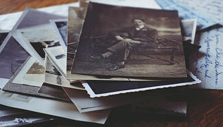 A stack of old photos and a handwritten page on a tabletop. The 1920s sepia-tone photo on top shows a seated man with a suit, bow tie and striped socks.