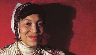 Black woman with freckles smiling, wearing a white cap with a black and red textured border. Her short black bangs extend from front of the cap. She wears a white turtleneck top with red blue and yellow jewels embroidered on the shoulders and a gold beaded necklace. The background is red and shows her profile in shadow