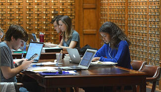 Two male and two female students sitting at a desk working on open laptops. Rows of wooden drawers of card catalog line the two walls behind them.