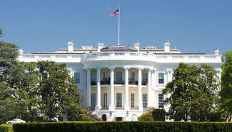 Photograph of the White House facade with green trees on either side and green hedge in front. An American flag flies above on long pole at center.