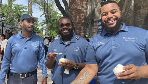 Three men wearing blue shirts that read "Yale Library Security" smile at camera. Man at far left wears a dark blue cap with white letter Y. Two men next to him hold plastic cups with vanilla ice cream.