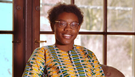 Student with short black curly hair wearing brown wire-rimmed glasses and a yellow, green, and white pattern shirt resembling an African textile sits smiling in front of doors with wooden panes of glass.
