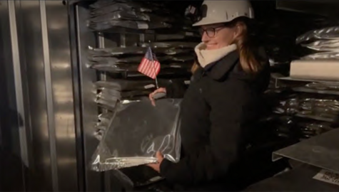 Woman with glasses and blonde hair, wearing jacket and white hard hat holds a foil wrapped flat package with the American flag on top.