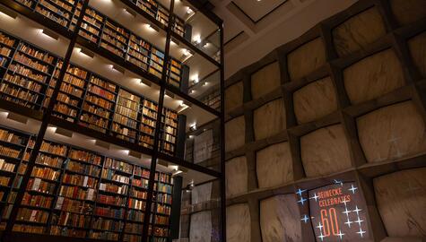 Interior view of Beinecke library of many stories of books on shelves behind glass across from marbled wall with projection that reads "Beinecke Celebrates 60"