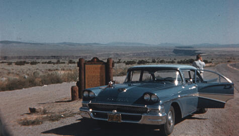 Desert scene showing horizon with wooden sign reading "Rio Grande" and a blue 1950s Chevrolet with open door and a man in short sleeve shirt with back to camera looking out at scene from back of the car.