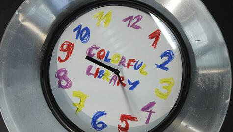 Clock face with wide silver rim with numbers with different-colored crayons and "Colorful Library" written in colorful crayons at center