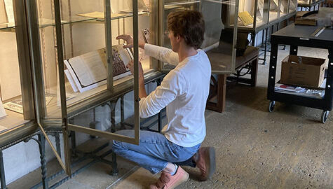 Student in white long-sleeve t-shirt, blue jeans, and pink sneakers squats before open display cases arranging a display of manuscript pages