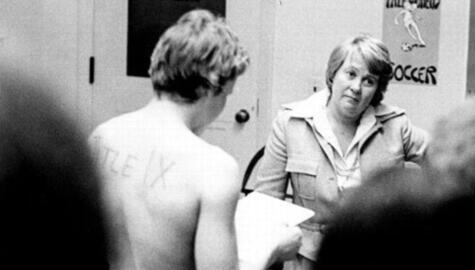 black and white photo shows shirtless woman with short hair and the words "Title IX" written on her back. She is facing away from camera, holding papers in her hand. To her right and looking off and to the right is a woman wearing a suit and open necked blouse. Blurred backs of heads of unidentified individuals are in the foreground and on the wall hangs a poster for Yale Women's Soccer.
