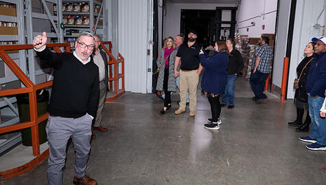 Man in dark sweater and grey pants and glasses gestures with right arm, leading tour in warehouse to group of 9 people who are gazing up.
