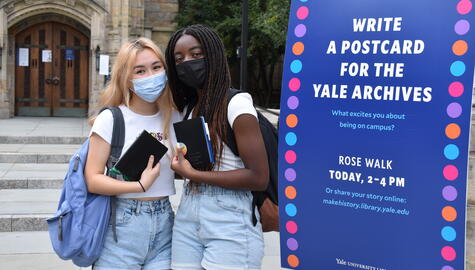 Two female students with library-branded journals standing next to the event posters