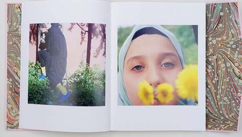 Inside book pages showing marbled papers inside front and back covers. Photo on left page shows woman in black Turkish dress with green socks and blue shoes stepping up a ladder in a garden with greenery around her feet; photo at right shows face of young girl looking at camera with three yellow flowers in the foreground in front of her mouth.