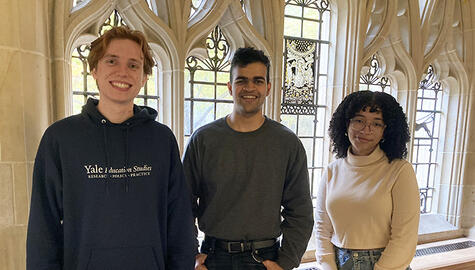 Three students pose before leaded glass gothic-style windows: man at far left has reddish hair and wears a blue hooded sweatshirt that reads in white lettering on front "Yale Education Studies"; the man at center has short dark hair and wears a round-neck shirt, black belt and black jeans and has his thumbs hooked into his front pockets; the short woman at right has curly black hair to her shoulders, wire-rimmed glasses, wearing a white turtle-neck and blue jeans
