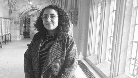 Woman with curly dark hair and wire-rimmed glasses poses alongside windows, with hallway and arched entrance behind her. The photo is black and white. She wears a striped turtleneck and plaid coat.