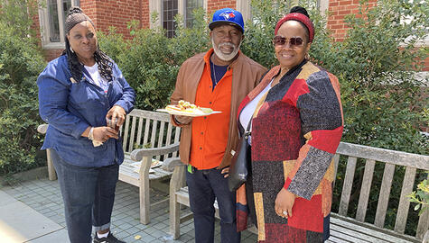 Two women and one man pose for camera. Woman at left wears blue shirt, man has blue cap and orange shirt and holds plate of food. Woman at far right wears multicolored sweater, sunglasses, and red hairband