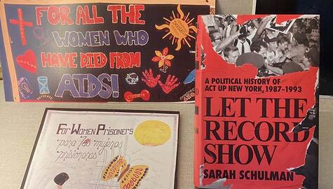 Closeup of a poster that reads "For all the women who have died from Aids!" and a framed white drawing that reads "For Women Prisoners" and the red cover of a book titled Let the Record Show: A Political History of Act Up New York 1987-1993