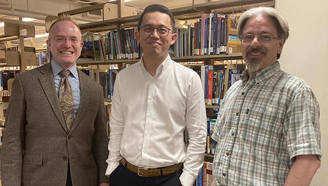 Three men pose in front of metal bookcases lined with books. Man at left has reddish hair and wire glasses and wears a brown suit and paisley tie;  man at center has black hair and dark rimmed glasses, white shirt, brown leather belt, and hands in pockets; man at right has grey hair and van dyke, with rimless glasses and short-sleeve blue, white and gray plaid shirt. All smile broadly.