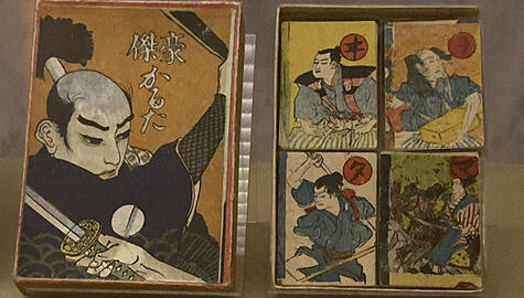 Large painted card on left shows bald Japanese warrior wielding sword. At right four smaller playing cards each show a Japanese man in traditional dress. Each card has a red circle with a Japanese character at center.