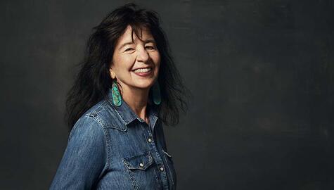 Woman with long black hair and bangs looks at camera smiling, She wears long oval turquoise earrings and a denim shirt open at the collar with front snap pockets