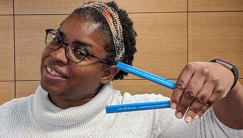 Smiling Black woman wearng white turtleneck sweater, glasses, and colorful hairband, holding two blue pencils printed with "catalog of US Government Publications" in white