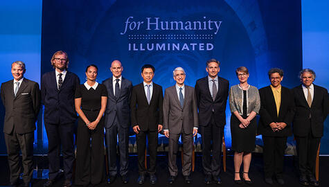 Ten standing people in dress clothing (8 men, 2 women) pose before blue background that displays type "For Humanity Illuminated"