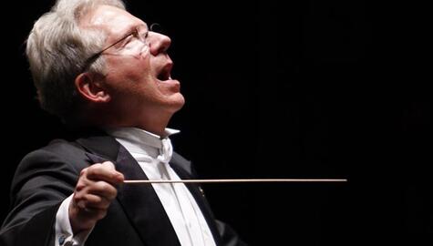 Man with white hair and wire-rim glasses, in black tuxedo with white shirt and cravatte, holds conductor's baton, leaning back with eyes closed and mouth open as if singing