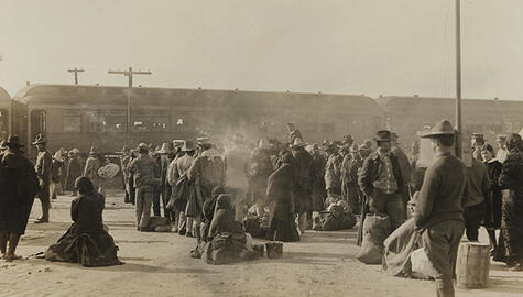 1914 sepia-tone photo showing crowds of people with suitcases, in a dusty setting, some seated some standing, with two train cars in the near distance