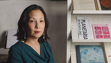 Woman with short dark hair and dark green blouse and long earrings looks at camera. To her left are shelves that display artwork. One reads "Planetaria by Monica Ong"