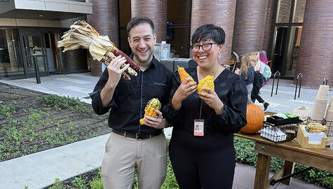 Smiling man and woman in black shirts stand together smiling holding yellow and orange gourds and dried corn stalks