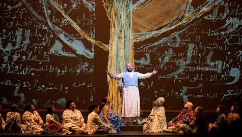Stage set with arabic script projected in white on black background. Man in Arabic dress stands with arms outstretched in front of animated image of a blue and yellow tree. Eleven people in long garments sit at his feet, six to his right and five to his left.