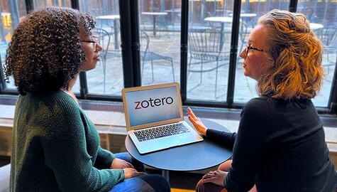 Young woman with short curly hair green sweater and glasses sits at small round desk with blond woman with dark shirt and glasses. On desk is a laptop with the word "Zotero" on the screen. Empty tables and chairs on patio are visible in background through glass wall. 