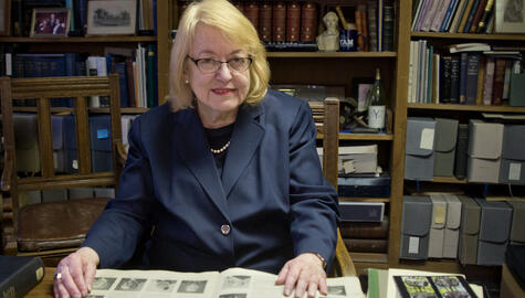 Seated woman with blonde hair in glasses wearing a navy suit with an open book in front of her and bookshelves  full of books, archival photo boxes, framed photos and other objects behind her