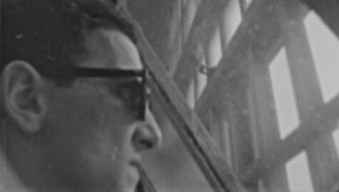 Black and white image of man in profile with dark glasses looks out panes of gridded concrete window