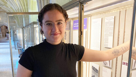 Curator Gabby Colangelo stands in front of open display case in Exhibition Corridor. She wears a short-sleeve black top, wire-rim glasses and long rectangular earrings. Her brown curly hair is tied back. She has a tattoo of a Greek vase with flowers on her forearm and reaches into the case, looking at the camera.