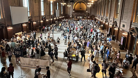large high-ceiling space filled with hundreds of students. At bottom right edge white sign on black standing frame reads "Welcome to Yale College Academic Fair"