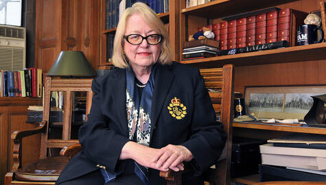 Portrait of Judy Schiff, who has chin-length blonde hair and wears dark-rimmed oval glasses, a navy sweater, scarf, and blazer, with a string of pearls at her neck. Her blazer has a red and gold Ralph Lauren insignia. She sits with one hand over the other, resting on the armrest of the chair. She sits in front of bookshelves.