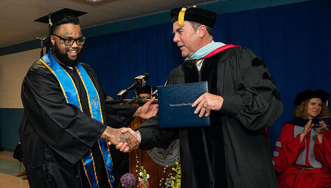Two men in black caps and gowns shaking hands. Student on left wears glasses and blue stole. Faculty member on right wears cap with gold tassel and holds blue leather diploma case.