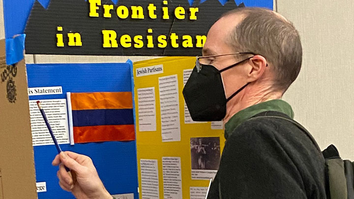 Man with light brown hair wearing glasses and black face mask and dark sweater and shirt points pencil at a panel of a display that is titled "Frontier in Resistance"