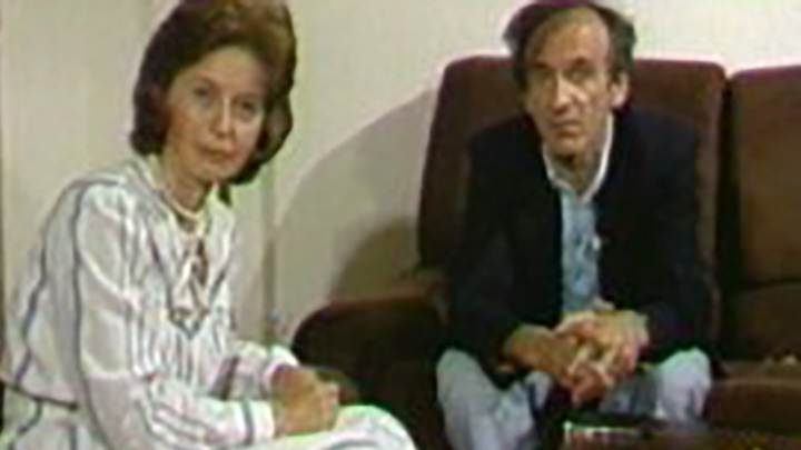 Woman in white dress with grey stripes looks at camera; next to her is seated man with dark jacket and hands folded