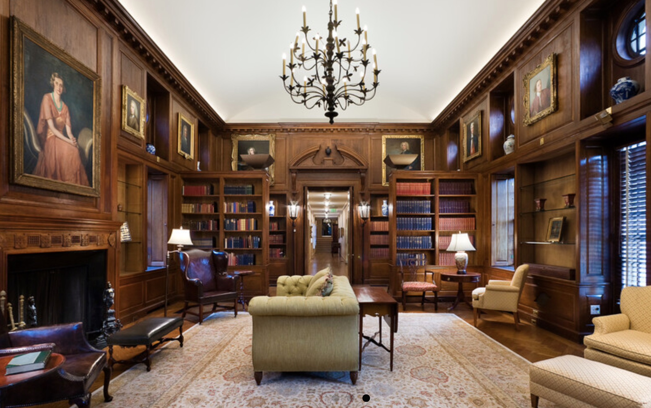 dark paneled high ceilinged library room lined with books cases and portraits and a sofa facing a fireplace.