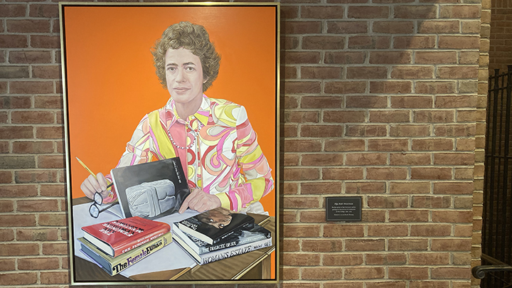 Painting of woman hangs on brick wall. She sits at a desk with six books on surface. She has short wavy brown hair and wears.a blouse with pink, orange, white, red swirls and holds black glasses and a pen in her right hand.