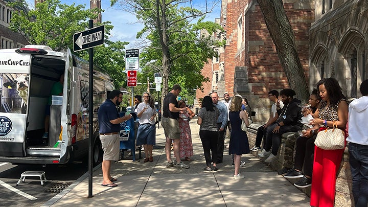 Street scene showing sidewalk full of standing people and people sitting on stone wall, talking together and eating ice cream. The white ice cream truck is at far left, with one of the rear doors swung open. Street sign in foreground reads "One way" with arrow and black type.