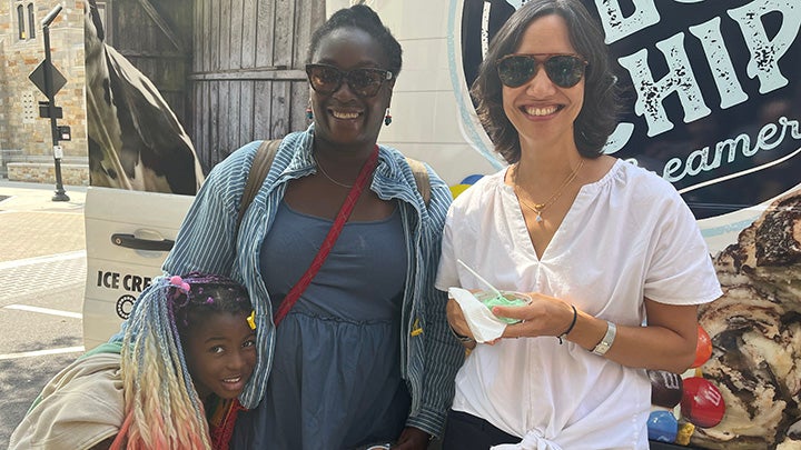 Small girl with long colorful braids hugs her mother who wears a blue dress and striped shirt and sunglasses. Next to her a smiling woman in white short-sleeve blouse and sunglasses holds a plastic cup with green ice cream and a spoon.