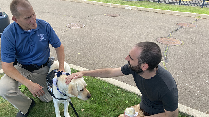 Man in dark tee shirt with black hair, beard and mustache pets Heidi the service dog, while talking with the dog's handler, wearing a Yale blue police force shirt and khaki pants. Heidi is wearing a black harness.