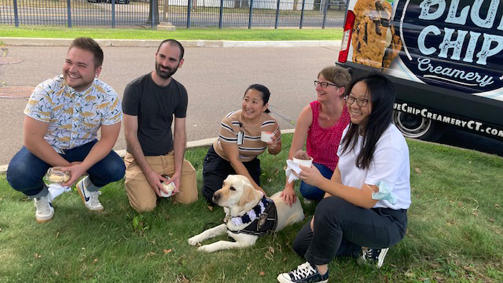 Five Winchester staff members sit on the grass with Heidi the labrador service ground between them. The man at far left wears a blue, white, and tan short-sleeve shirt and white Nike sneakers. The man next to him has a dark beard and mustache, a black tee shirt, and light brown pants. The woman next to him has a brown and black striped shirt and black pants. The woman next to her has short brown hair, glasses, and a red sleeveless scoop-neck top. The next woman has a white blouse, dark pants, and black hair