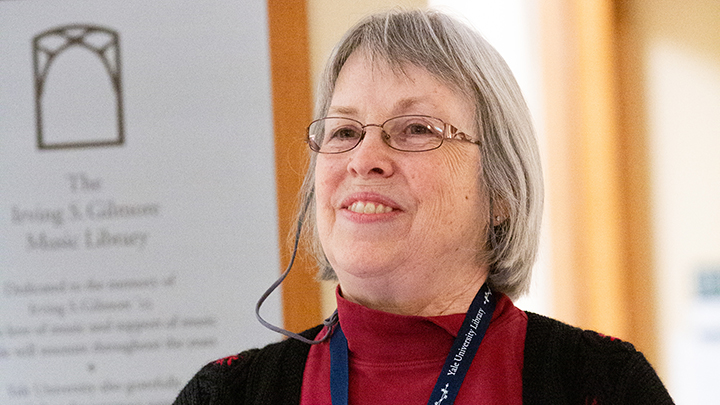 Smiling woman with short white hair, brown rim glasses, maroon turtleneck and black sweater. She wears a navy lanyard with white words "Yale University Library"