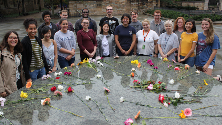 Two rows of students and staff standing behind a stone surface strewn with cut flowers.